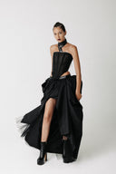A model wearing a black strapless corset with a high-low draped Sisi skirt, and black platform ankle boots, posed against a plain light gray studio background.
