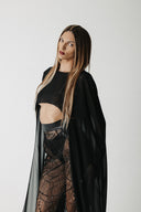A woman modeling a black gothic-style outfit featuring a sheer black cape crop top, and a black lace skirt, set against a plain light gray studio background.
