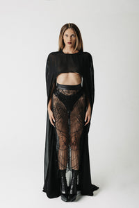 A model stands against a light gray studio background wearing a black sheer long-sleeve top with a cape-like drape, black lace high-waisted bottoms, and black ankle boots.
