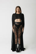 A model stands against a light gray studio background wearing a black sheer long-sleeve top with a cape-like drape, black lace high-waisted bottoms, and black ankle boots.