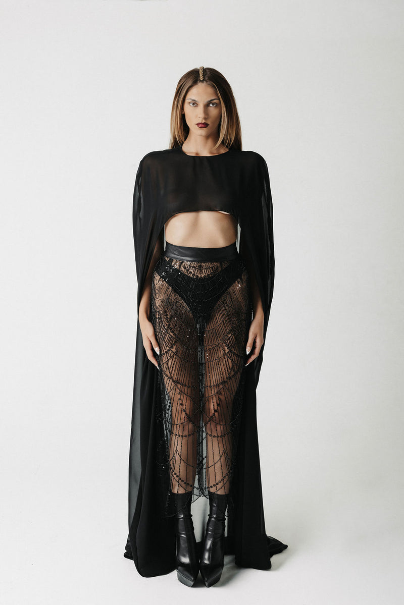 A model stands against a light gray studio background wearing a black sheer long-sleeve top with a cape-like drape, black lace high-waisted bottoms, and black ankle boots.